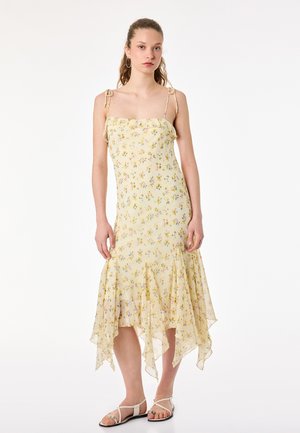 Ung kvinde iført en gul midikjole med blomsterprint, tynde stropper og asymmetrisk kant, kombineret med hvide flade sandaler, stående mod en hvid baggrund.