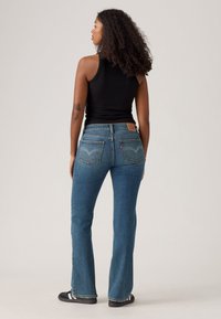 Femme aux cheveux bouclés portant un haut sans manches noir, un jean en denim bleu et des baskets noires, se tenant de dos contre un fond uni.