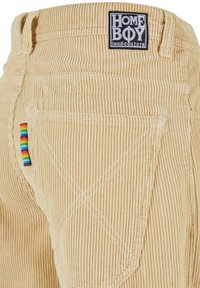 Pantaloni in corduroy beige con etichetta in vita, cuciture bianche e un accento a strisce arcobaleno sulla tasca posteriore.