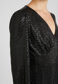 Černé šaty s hlubokým výstřihem do V, které mají strukturovanou látku zdobenou stříbrnými flitry, dlouhé rukávy a přiléhavý střih.