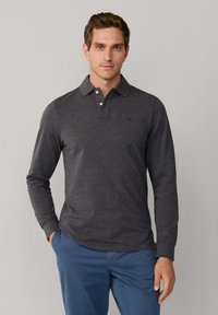 Polo gris à manches longues avec un col boutonné, tissu texturé et un logo discret sur la poitrine. Porté avec un pantalon bleu.