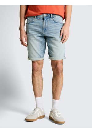 Mężczyzna w jasnoniebieskich podwiniętych denimowych szortach, białych skarpetkach i beżowych tenisówkach, stojący na jasnym, jednolitym tle.