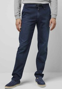 Pantalon droit bleu foncé en denim avec une taille à cordon de serrage ajustable. Porté avec des chaussures slip-on navy et un sweatshirt gris.