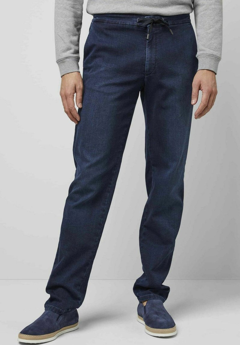 Pantalon droit bleu foncé en denim avec une taille à cordon de serrage ajustable. Porté avec des chaussures slip-on navy et un sweatshirt gris.