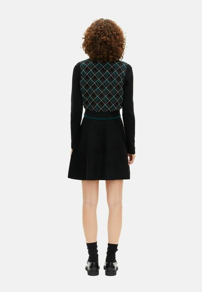 Mulher com cabelo encaracolado, vestindo um vestido preto com um padrão quadriculado verde e azul na parte de trás, meias pretas e sapatos pretos, em pé.