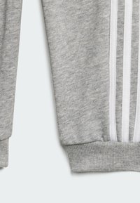 Manica di felpa grigia con polsino a costine. Presenta tre strisce bianche verticali lungo il lato. Tessuto morbido, design casual.