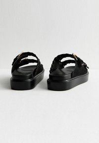 Sandales noires à semelle plate, avec des brides tressées et des accents en métal doré. Texture lisse avec un design minimaliste.