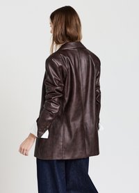 Femme portant un blazer en cuir marron foncé avec des manches froncées sur une chemise blanche et un pantalon large bleu marine, de dos par rapport à l'appareil photo.