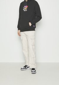 Personne portant un sweat à capuche noir avec un motif de dessin animé, un pantalon cargo beige avec des poches et des baskets noires et blanches, debout contre un mur blanc.
