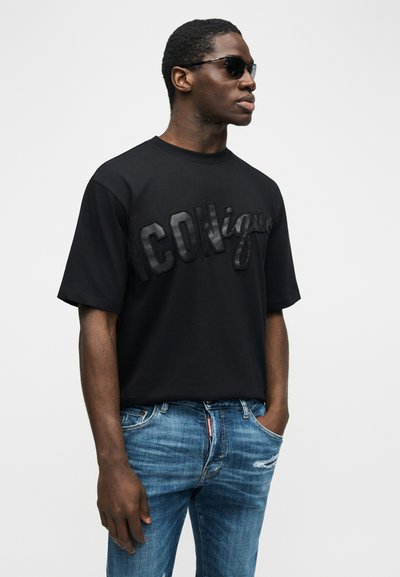 Άνδρας που φοράει μαύρα γυαλιά ηλίου, μια μαύρη φαρδιά μπλούζα με το κείμενο "ICONique" και μπλε τζιν με σκισίματα, στέκεται με τα χέρια στις τσέπες.