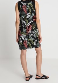Black sleeveless dress with a tropical leaf pattern in green, white, and pink. Model wearing flat black sandals. Smooth fabric texture.