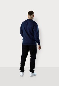 Navy blue sweatshirt featuring a crew neck and long sleeves, paired with black pants and white sneakers. Back view with tattoo visible.