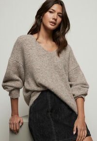 Pull en maille beige avec un large col en V et des manches surdimensionnées, superposé sur une jupe noire avec des détails de coutures visibles.