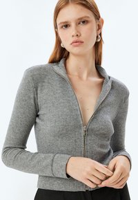 Cardigan grigio a coste con zip e colletto alto, maniche lunghe. Presenta una zip argento e un design aderente. Texture liscia.