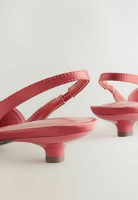 Des sandales ouvertes en satin rose arborent de fines brides autour de la cheville, un petit talon rose brillant et une surface lisse avec de subtils détails de couture.