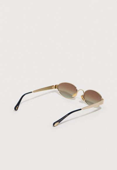 Στρογγυλά γυαλιά ηλίου με μεταλλικό χρυσό πλαίσιο, καφέ φακούς με διαβάθμιση και μαύρες άκρες στα πλαϊνά, πάνω σε απλή λευκή επιφάνεια.