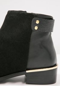 Bottine noire à la cheville avec textures mixtes en daim et cuir, clous dorés et talon bas empilé avec accent métallique.