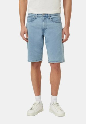 Mężczyzna ubrany w jasnoniebieskie szorty jeansowe, białe skarpetki i białe trampki, stojący na jednolitym tle.