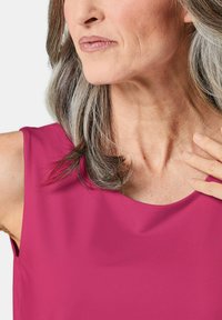 Top magenta senza maniche con una texture liscia e un collo tondo; il modello ha i capelli argento fino alle spalle e dettagli della pelle lievemente visibili.