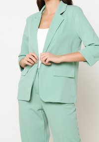 Miętowy garnitur z dopasowaną marynarką i pasującymi spodniami. Marynarka posiada klapę i dwie przednie kieszenie. Gładka tekstura materiału.
