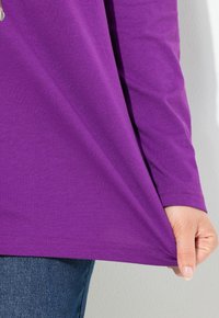 Lila langärmliges Hemd mit glatter Textur und geradem Saum. Die Hand zieht leicht am unteren Rand.