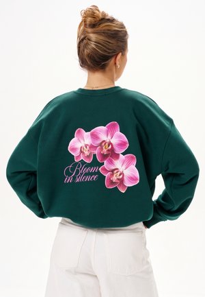 Vrouw met blond haar in een knot, draagt een donkergroene trui met roze orchideeënbloemen en de tekst "Bloom in silence" op de rug.