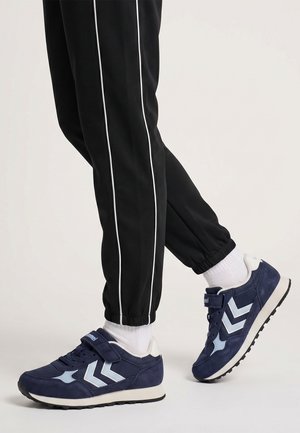 Nogi w czarnych spodniach z białymi paskami i białych skarpetkach w niebiesko-białe sneakersy na rzepy, stojące na jasnym tle.