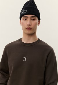 Joven con un gorro de punto negro con letras y una sudadera marrón oscuro que presenta un pequeño número romano II blanco en el pecho.