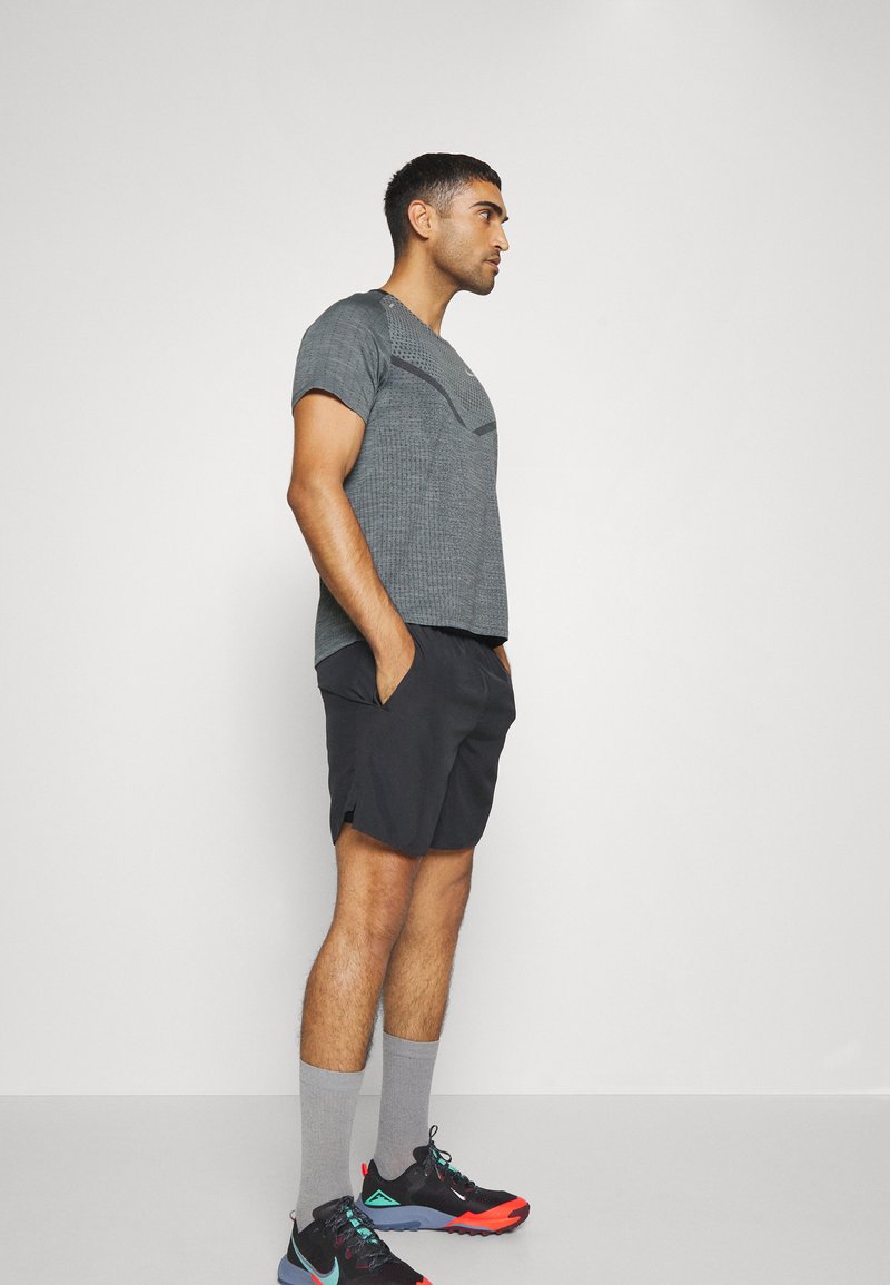 Grau strukturiertes Kurzarmhemd, schwarze Shorts, graue Socken, schwarz-rote Sportschuhe. Seitenansicht mit Händen in den Taschen.