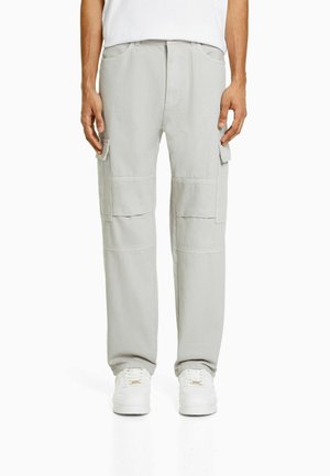 Homme portant un pantalon cargo gris clair avec des poches à rabat et des baskets blanches, debout devant un fond blanc uni.