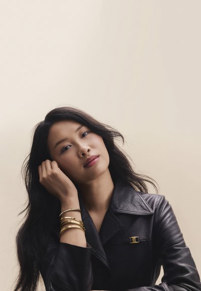 Femme aux longs cheveux noirs portant une veste en cuir noire et des bracelets dorés, reposant sa tête sur sa main sur un fond uni.