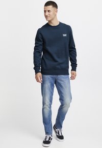 Donkerblauwe sweatshirt met ronde hals, geribbelde manchetten en onderrand; gecombineerd met lichtblauwe spijkerbroek en zwarte sneakers met witte accenten.