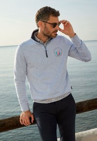 Lichtblauwe pullover met een ritskraag, voorzien van een kleurrijk zeilbootlogo. De binnenkant van de kraag toont een bloemenpatroon. Gecombineerd met donkere broek.