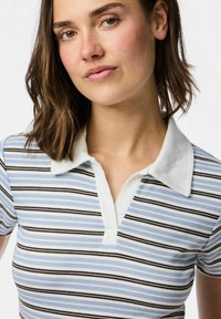 Pola krekls ar balto apkakli un zilām, melnām un baltām svītrām. Ribu tekstūra, īsām piedurknēm un pogu slēgšanas detaļa.