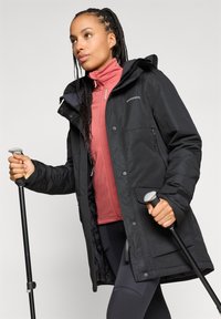 Czarna wodoodporna kurtka z kapturem, wyposażona w kieszenie na zamek i mankiety na rzepy, noszona na różowej warstwie środkowej i czarnych legginsach.