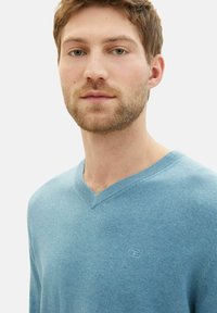 Hellblaues V-Ausschnitt-Pullover aus weichem Material, mit gerippten Bündchen und Saum, und einem kleinen Logo, das auf der Brust gestickt ist.