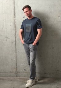 Námořnická modrá tričko s grafickým designem, světle šedé džíny a krémové tenisky. Stojí proti texturované betonové zdi.