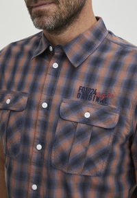 Camisa de botones con estampado de cuadros en tonos naranja y azul marino, con dos bolsillos en el pecho y detalle de logo bordado en el lado izquierdo.