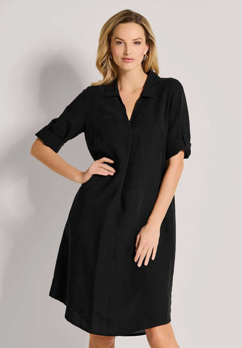 Schwarzes Hemdkleid mit Kragen, knielang, kurzen umgeschlagenen Ärmeln und lockerer Passform. Hergestellt aus einem glatten, leichten Stoff.