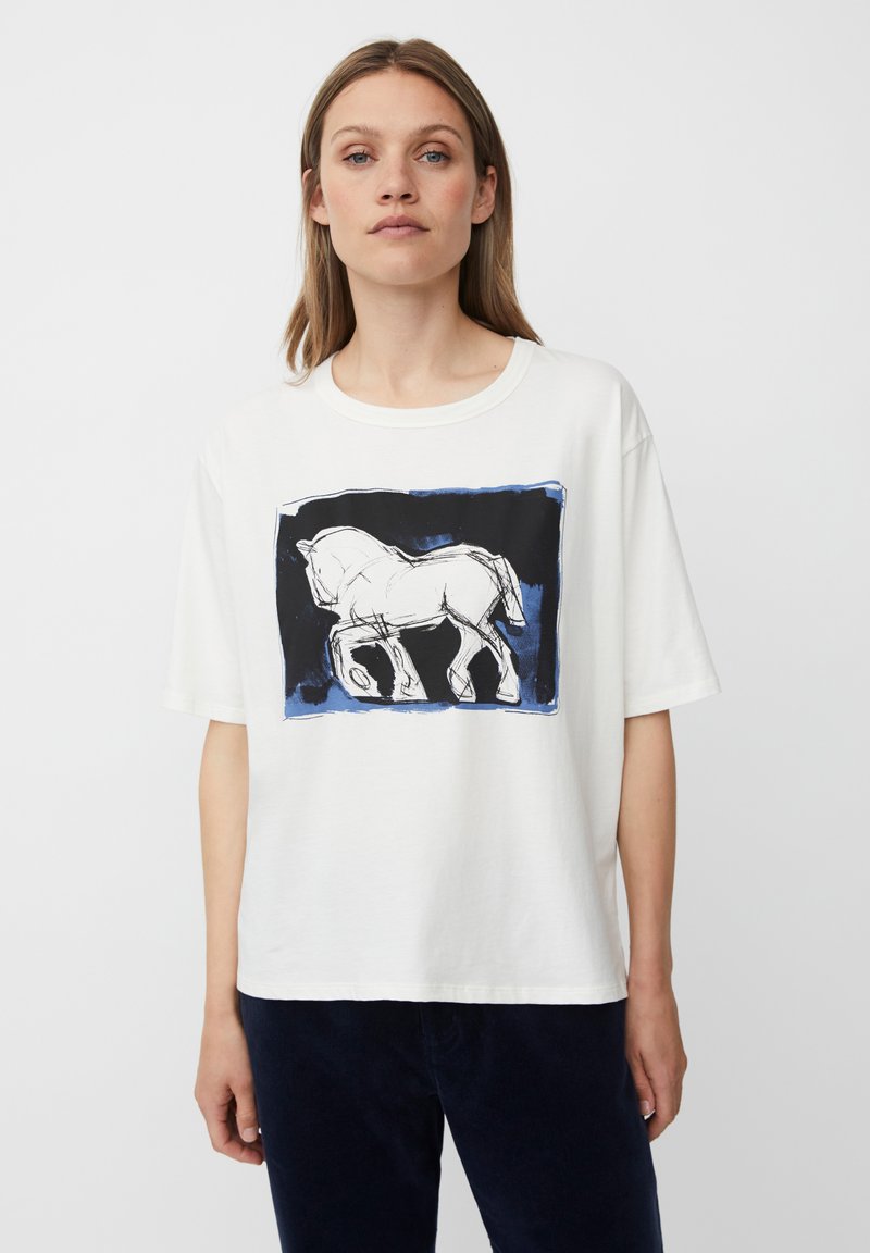 Frau trägt ein weißes T-Shirt mit abstraktem Schwarz-Weiß-Pferdedesign auf blauem und schwarzem rechteckigen Hintergrund, dunkle Hose.
