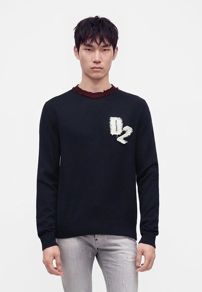 Jeune homme portant un pull bleu foncé avec un patch "D2" effiloché sur la poitrine et un jean gris clair usé, debout sur un fond uni.