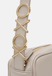 Tracolla in finta pelle beige con hardware dorato caratterizzata da disegni circolari e a croce, attaccata a una borsa rettangolare con una zip dorata.