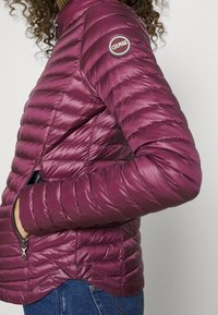 Vinröd puffjacka med kviltat mönster, dragkedjor på fickor och en rund logopatch på ärmen. Lättviktig och strömlinjeformad konstruktion.