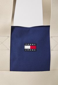 Blå tygväska med beige handtag och rektangulär Tommy Jeans-logotyp i vitt, rött och svart centrerad på väskan.