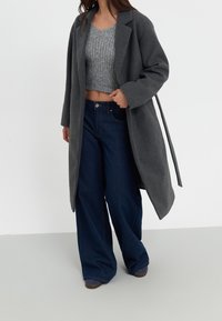 Šedý vlněný kabát s klopami a páskem, nošený přes světle šedý zkrácený svetr a široké tmavě modré džíny, vše na neutrálním pozadí.