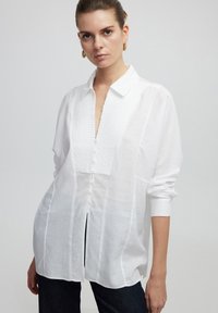 Camicia bianca a bottoni con pannello frontale testurizzato, maniche lunghe e colletto. Presenta spacco laterale e una vestibilità rilassata. Tessuto morbido.
