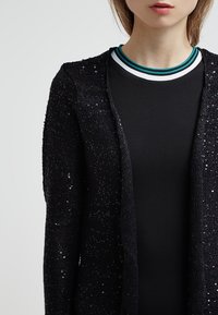 Svart cardigan med en texturerad stickad design, prydd med paljetter. Under den finns en svart topp med en randig krage. Enkel och modern.