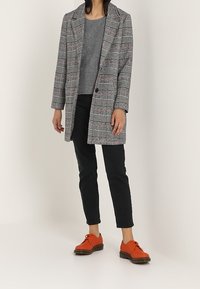 Cappotto a quadri grigio e bianco con accenti rossi, maglione grigio, jeans neri e scarpe arancione brillante con lacci e suola spessa.