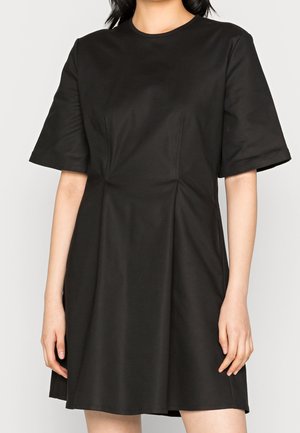 Kobieta ubrana w czarną sukienkę do kolan z krótkimi rękawami i okrągłym dekoltem, stojąca z ramionami swobodnie opuszczonymi wzdłuż ciała.