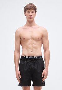 Μαύρο μαγιό σορτς με ελαστική μέση που φέρει το λευκό λογότυπο 'CALVIN KLEIN', κατασκευασμένο από λεία, ελαφριά υλικά.